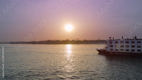 A luxury cruise ship sailing on the Nile River in Aswan, Egypt, during a beautiful golden sunset with a hazy sky.