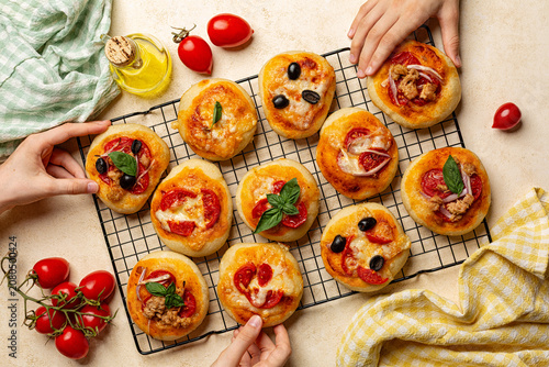 Family sharing food and children hands reaching for the pizza. Homemade mini pizzas tomato sauce, mozzarella, cherry tomatoes, basil,  tuna, onion toppings, served on a cooling rack. Directly above.