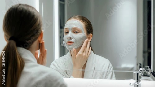 Young woman applying a clay face mask in a bathroom mirror. Skincare routine and beauty treatment at home or spa. Wellness and rest concept