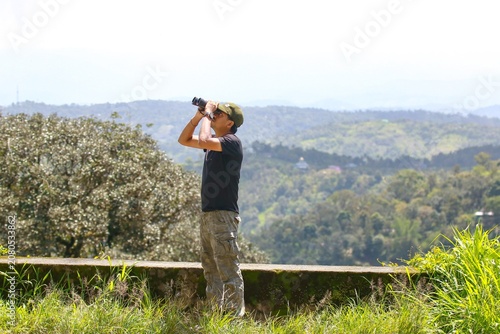 Male birdwatcher using binoculars at a panoramic viewpoint in the Western Ghats of Munnar, India, searching for birds across the landscape