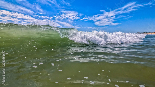 close up view of an ocean wave coming into shore with a bright blue sky and clouds in the background and the beach way off in the distance