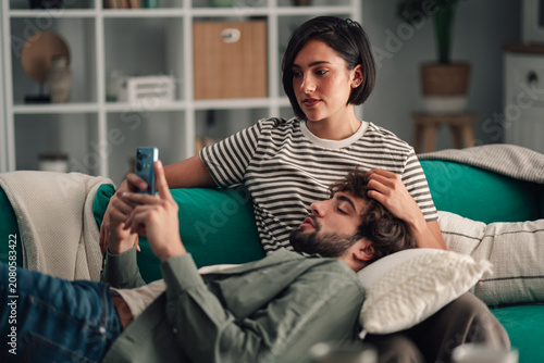 Fotografie Young couple relaxing together on sofa, man holding smartphone while woman cares