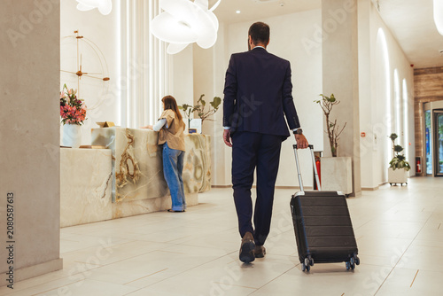 Business Traveler Pulling Carry-On Suitcase Through Modern Hotel Lobby Toward Reception Desk