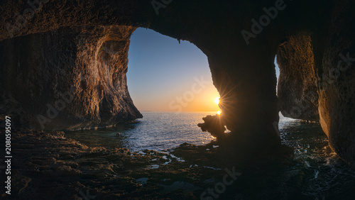 Dark rocky cave frames a golden sunrise over the calm sea along the Golfo di Orosei coast.
