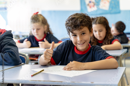 Smiling elementary school boy student showing thumbs up gesture, sitting at desk in a bright classroom with classmates