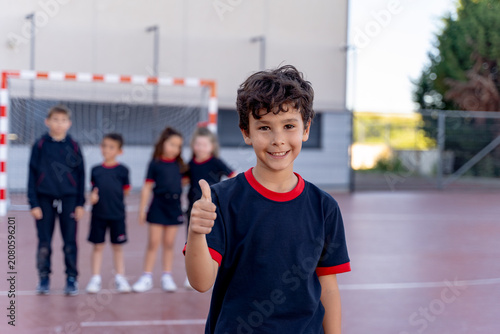 Young boy smiling and showing a thumbs up gesture, standing with schoolmates on a sports court after physical education class