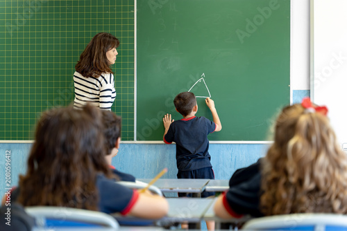 Teacher guiding a young student drawing a triangle on the classroom chalkboard, other children watching from their desks