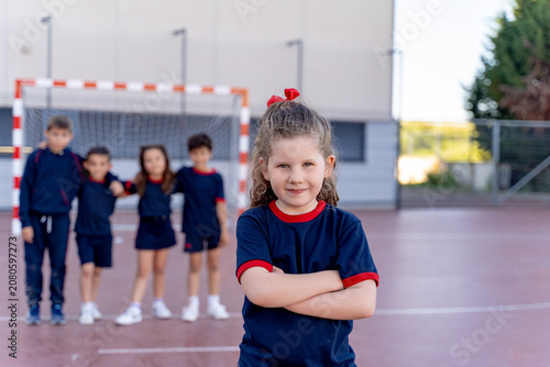 Confident young girl in school uniform smiling, arms crossed, with blurred classmates standing by a goal in a sports court