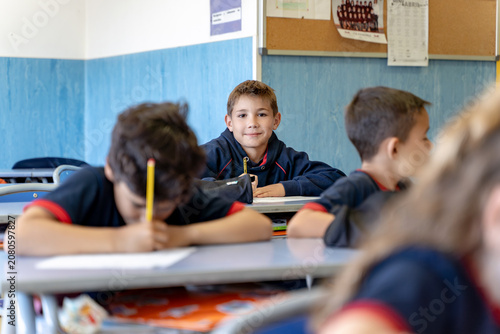 Happy elementary student sitting at a desk, looking at the camera and smiling, with classmates working in front