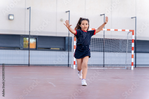 Girl running with arms outstretched on a school playground, wearing sportswear and celebrating a goal on the court