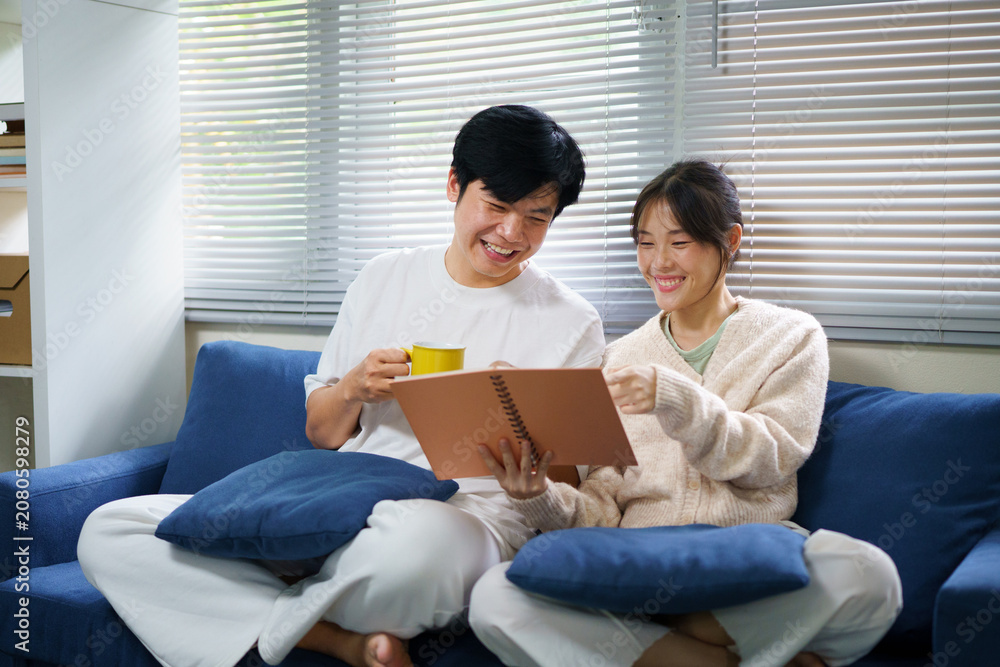 Fototapeta premium Young happy couple sits together on a cozy blue couch in their living room, looking at a memory book together and smiling while reminiscing about cherished times and drinking coffee.