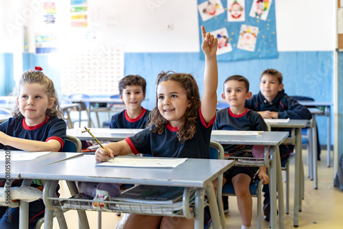 Young girl raising hand in a classroom with other smiling students, showing active learning and engagement in education