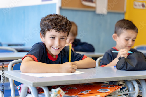 Young student smiling at camera while sitting at his desk, writing on paper in a classroom setting