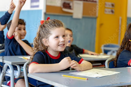 Young female student smiling while actively participating in a school lesson alongside classmates, raising hands