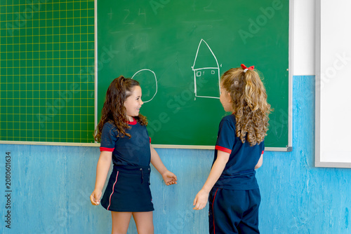 Young girls in school uniform looking at each other, drawing shapes on a blackboard during a class creating friendship and learning