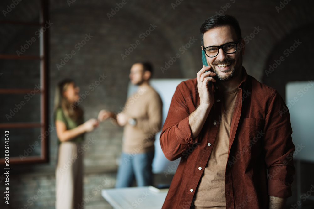 © Novak - A confident businessman standing or sitting at his desk, talking on a smartphone, while his professional colleagues work and collaborate in the blurred background. © Novak - A confident businessman standing or sitting at his desk, talking on a smartphone, while his professional colleagues work and collaborate in the blurred background.