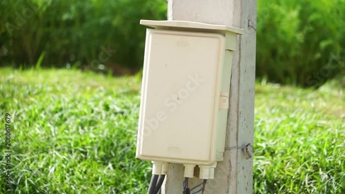 A box is mounted on a utility pole, cables connect to the box and restt on the ground, the area has green grass and some plants, It is bight outside, indicationg daytiame.