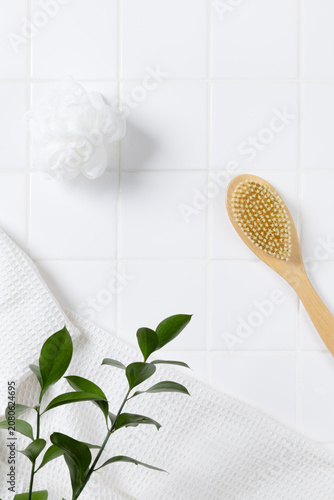 Top view of organic spa product mockup background with green leaves. Flat lay of body brush, bath puff, and white towel on square tile wall with copy space for cosmetic display.
