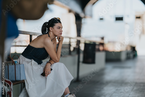 A young tourist woman sits on a bench at a train station, holding her phone and luggage. She waits thoughtfully on the covered platform, appearing reflective and patient.