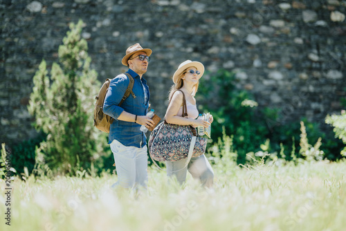 Foto Tourists: a man and woman walk together carrying bags and water on a sunny day