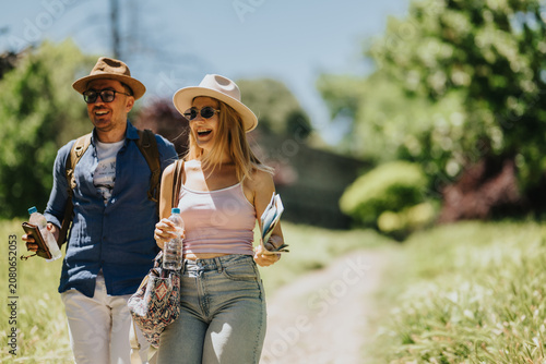 A smiling couple of tourists walk along a sunlit trail carrying backpacks  water bottles and a map. The pair enjoy a relaxed summer walk outdoors while sightseeing and exploring nature.