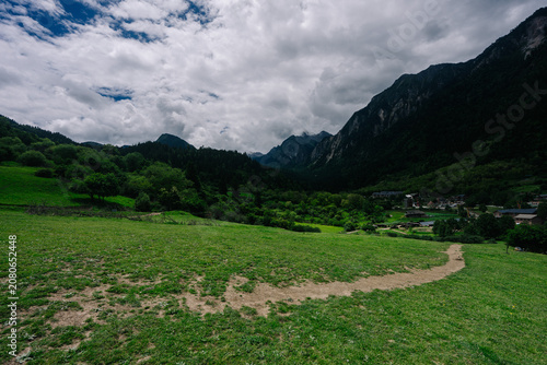 A serene valley landscape with lush green fields, a winding path, and towering mountains under a cloudy sky.