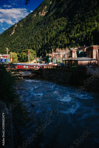 A river flows beneath a bridge in a mountainous landscape, with buildings and a cloudy sky visible in the background.