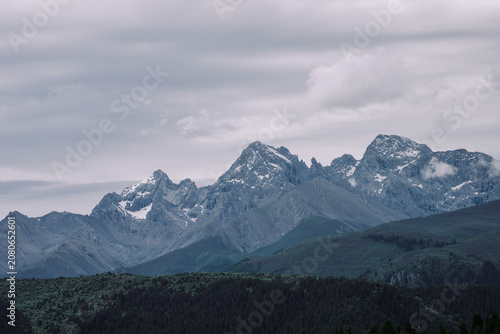 Dramatic mountain range shrouded in mist and clouds, with snow-capped peaks and a dark, forested foreground.