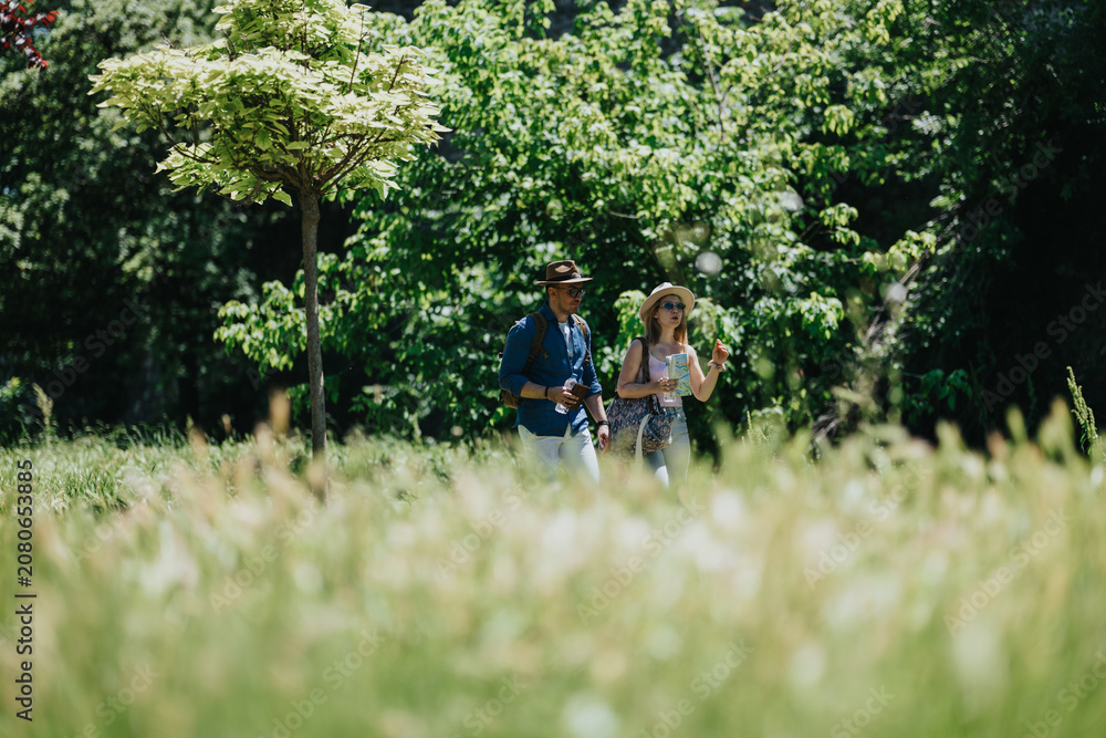 Fototapeta premium A couple of tourists walk through a sunlit park holding a map and drinking water. The pair stroll together in a leafy green setting while exploring outdoors.