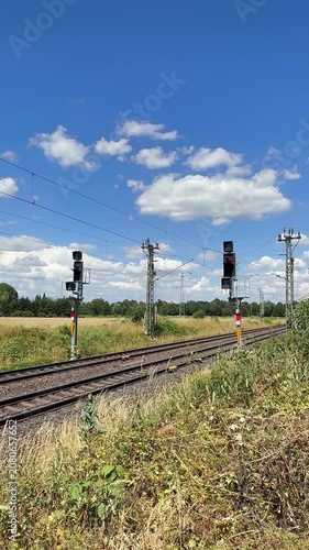 Bahnstrecke mit Signalen unter blauem Sommerhimmel | Hochformat - Ruhige Bahnstrecke mit Eisenbahnsignalen, Gleisen und sommerlicher Landschaft unter wolkenreichem Himmel. Ideal für Mobilität, Infrast