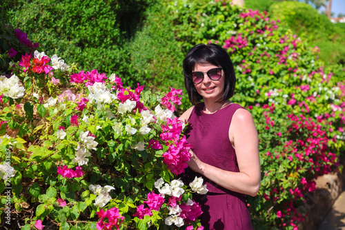 Elegant 40-year-old woman in beautiful dress posing near vibrant flower bed in sunny garden.