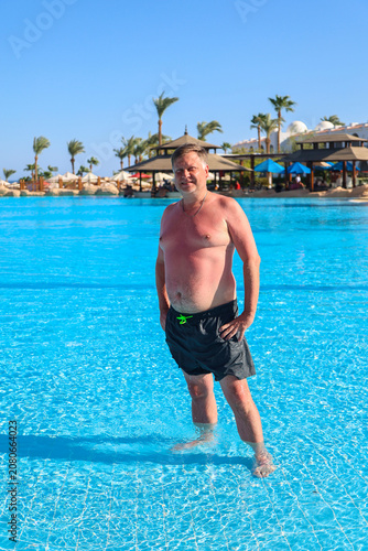 Tanned adult blond European man standing in a resort hotel pool under bright sunny skies.