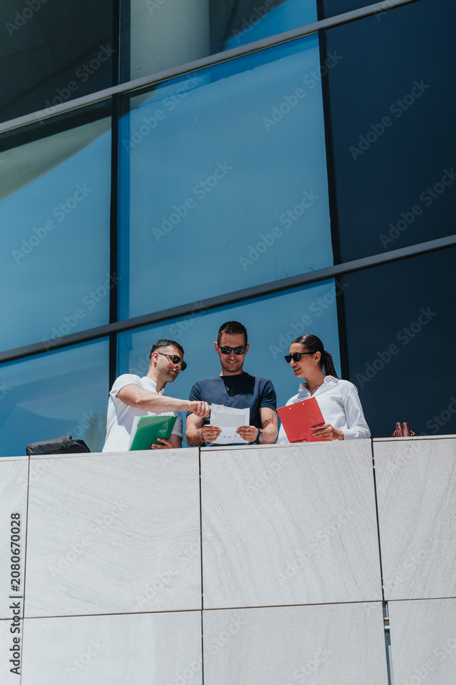Fototapeta premium Three business associates examine paperwork together while standing on an office balcony. Colleagues collaborate and discuss documents in front of a modern glass building.