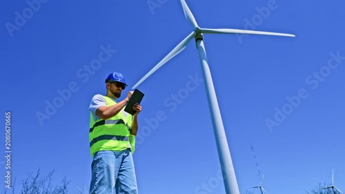 A windmill engineer takes notes on a tablet while inspecting a wind turbine at a wind farm. The sky is clear and blue as he focuses on his task in the field.