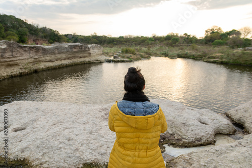 Fotografía Woman in Yellow Winter Jacket Looking at River from Rocky Shore at Sunset
