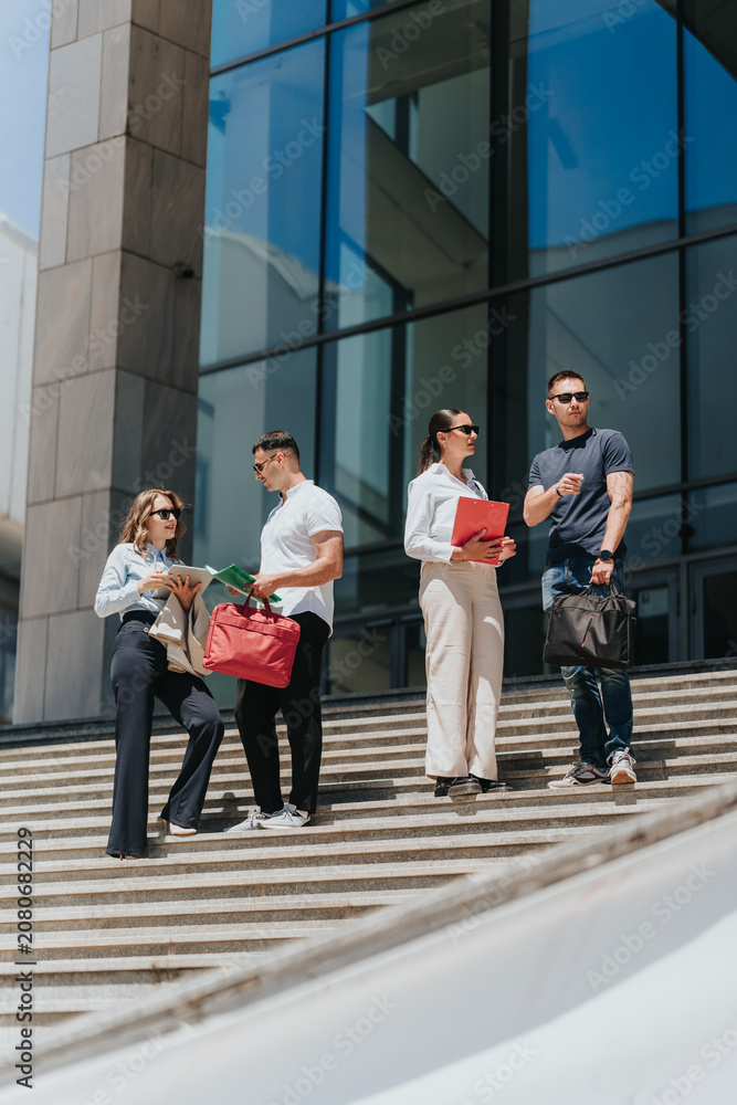 Fototapeta premium Four business associates stand on outdoor steps holding folders, bags and talking. Colleagues converse on the steps of a modern office building while reviewing documents.