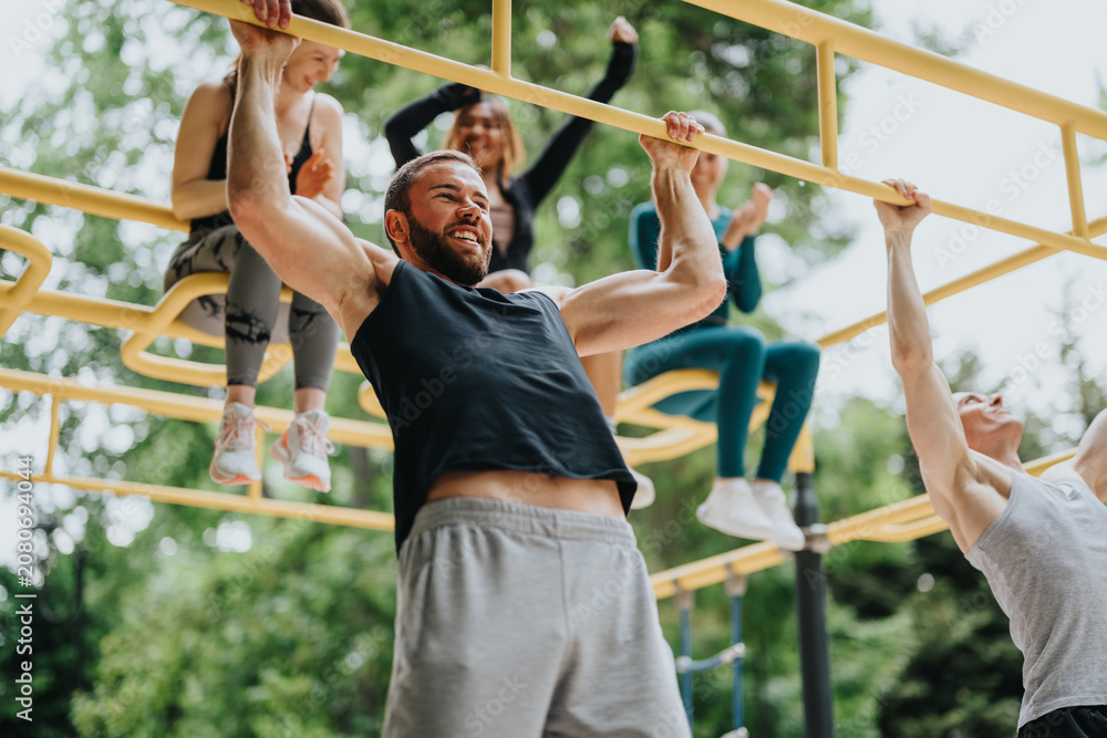 Fototapeta premium A muscular man performs a pull-up on yellow bars while friends cheer and sit above him. Casual outdoor gym workout shows a group exercising together and celebrating fitness.