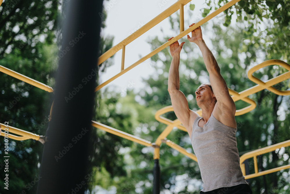 Fototapeta premium A muscular man grips yellow monkey bars while exercising outdoors. He trains on playground-style bars during an outdoor workout session.