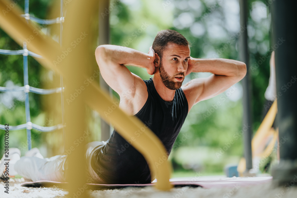 Fototapeta premium A fit young man performs abdominal crunches on a mat with his hands behind his head. He trains core muscles outdoors in a focused workout session.