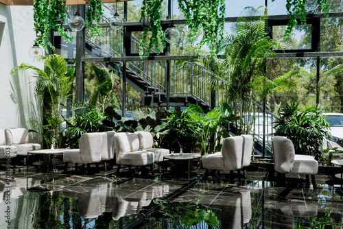 Contemporary glass house conservatory restaurant seating area equipped with premium furniture, hanging ivy, and architectural steel staircase background