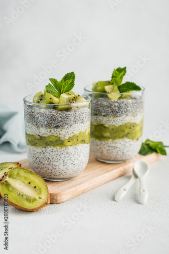 Two glasses of layered chia seed pudding with milk, kiwi puree, and fresh kiwi slices, decorated with mint leaves on a wooden board