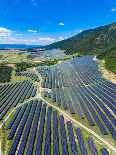 Solar panels cover large fields under a blue sky with scattered clouds in a rural area near mountains on an sunny day