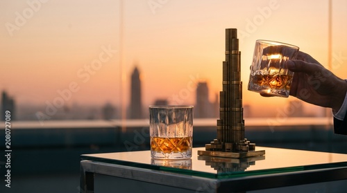A hand holds a glass of amber whiskey against a vibrant sunset over a city skyline, with a miniature skyscraper model nearby.