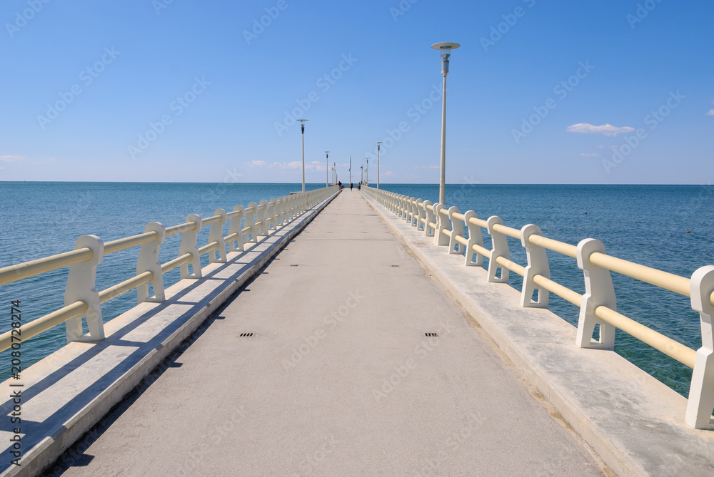 Fototapeta premium Long concrete pier extending into a calm blue sea beneath a clear sky, featuring strong leading lines, symmetry, and a peaceful coastal atmosphere with ample copy space.