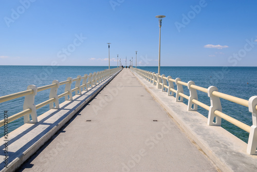 Long concrete pier extending into a calm blue sea beneath a clear sky, featuring strong leading lines, symmetry, and a peaceful coastal atmosphere with ample copy space.