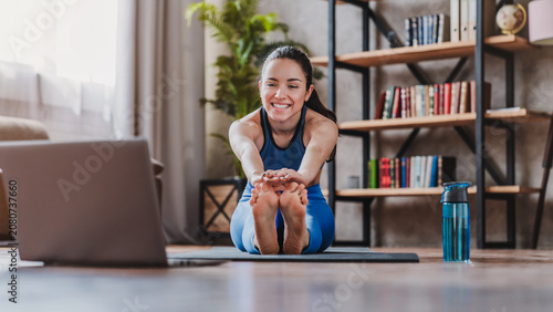 Sporty young woman stretching on mat while watching online tutorial on laptop at home, fit female exercising indoors, home workout, fitness and healthy lifestyle concept
