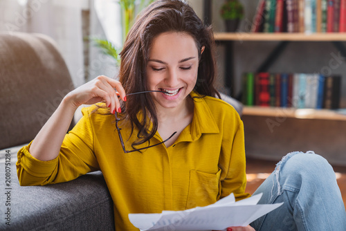 Young smiling woman sitting on floor and checking bills in living room, happy female managing household budget, financial planning and home expenses concept