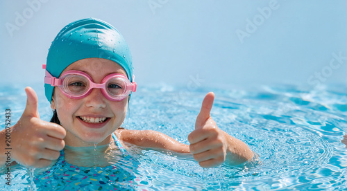Happy young girl wearing a blue swimming cap and pink goggles giving a double thumbs up while playing in a sunny outdoor pool