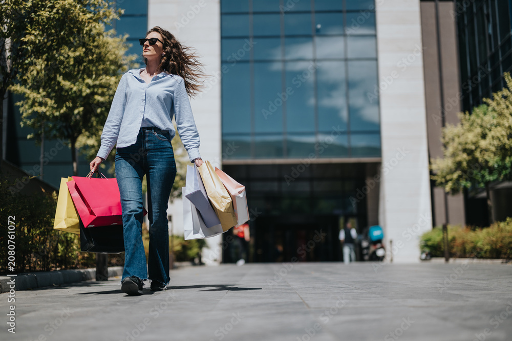 Fototapeta premium A woman walks confidently with multiple shopping bags in front of a shopping mall. The shopper strolls past the mall entrance on a bright sunny day, holding colorful bags.