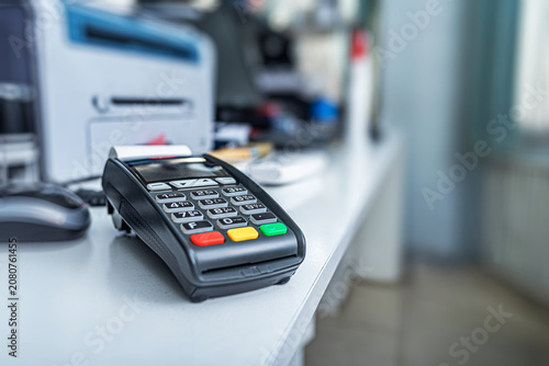 A black credit card terminal sits on a white office desk next to a computer mouse and a printer.